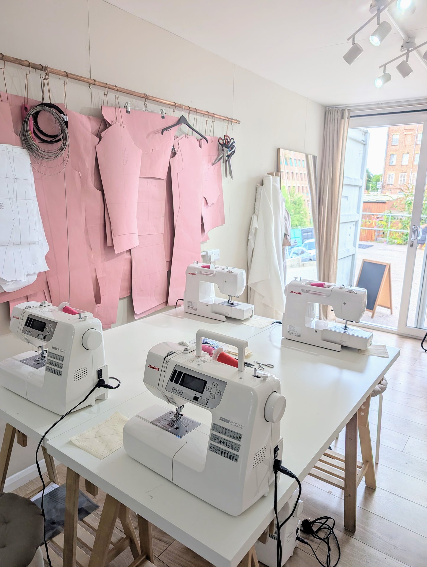 Sewing room with multiple sewing machines on a table, pink fabric in the background.